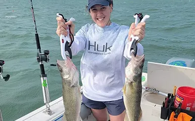 A cheerful female angler on a More Gooder Fishing Charters boat holding two large Walleye fish, with Lake Erie's waters in the background, dated June 7, 2022.