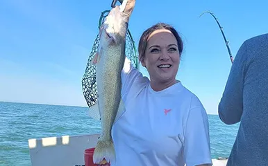 A female angler holding a caught Walleye with a bent fishing rod in the background on a More Gooder Fishing Charters boat on Lake Erie, June 10, 2022.