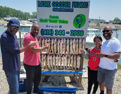 A joyful group of four, including a child, proudly presenting their fishing haul on a stand, with the More Gooder Fishing Charters sign and the calm marina backdrop on a bright day, June 9, 2023.