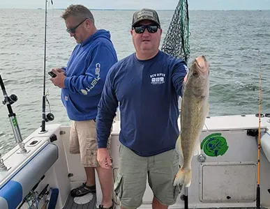 Two men on a More Gooder Fishing Charters boat, one holding a large walleye, with fishing gear on deck and overcast skies overhead, embodying the successful and serene fishing experience.